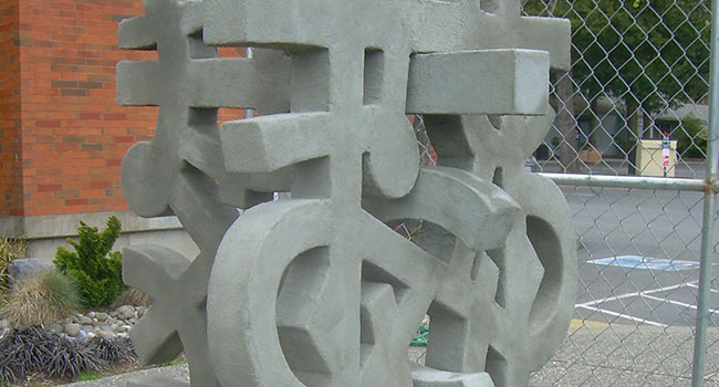 Close up of a concrete outdoor sculpture. The sculpture is gray with blocky linear forms that cross and merge. Most forms are straight but some make curved shapes, inclding an infinity sign. In the background is a brick wall and a parking lot.