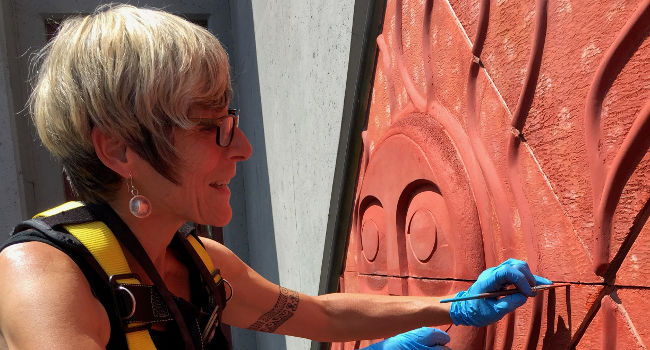 A woman wearing a safety harness and blue gloves as she performs conservation treatment on a low relief sculpture on the side of a building.