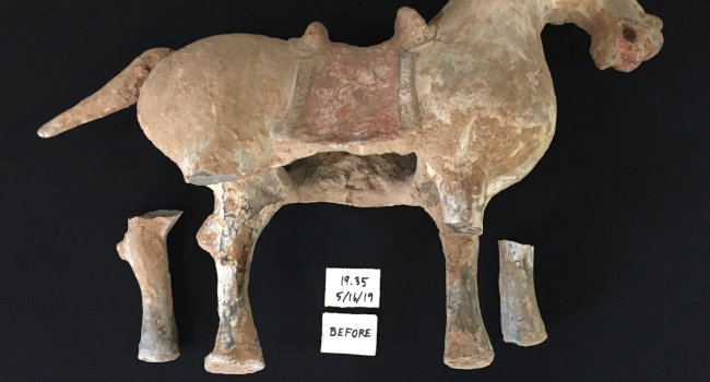A terra cotta horse with two legs broken off. The horse is laid on its side showing a cavity in the belly. On either side of the horse's intact legs is one of the broken off legs. There is a label between the horse's legs with the word 