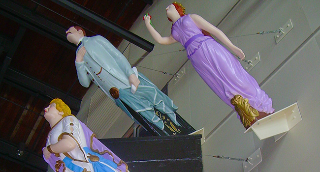 Three ship figureheads mounted on a wall. A male figurehead wears a blue suit. A red-haired female figurehead wears a purple dress. A yellow-haired female figurehead wears a dress in dark and light purple and blue with gold trim and ornaments.
