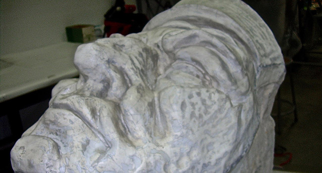 A section of a large terra cotta sculpture before conservation treatment. The section is half of the face of a figure. The gray surface is unevenly discolored.