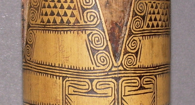 Close up of a cylindrical container made of light yellow wood with black line decorations. The compartmentalized design uses narrow parallel points, whorls and small triangles.