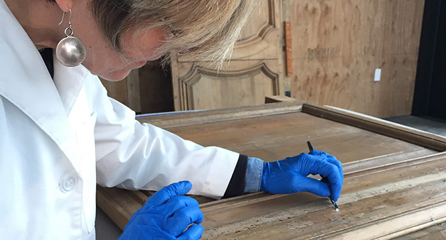 A woman wearing a lab coat and blue nitrile gloves holding a small metal spatula while working on a large carved wooden panel.