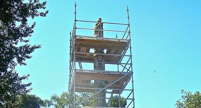 Outdoor scene looking up at the top of a totem pole surrounded by scaffolding. The sky is blue and clear and there are several trees in the perimeter of the image.