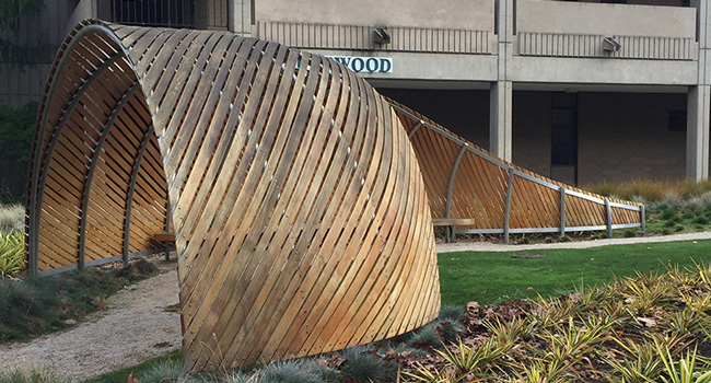 A large outdoor sculpture built from thin metal supports and diagonally-laid long pieces of wood. A pathway leads into the large arched end of the sculpture on the left and another path is visible near the narrow end. A building is in the background and there are plants around the sculpture.