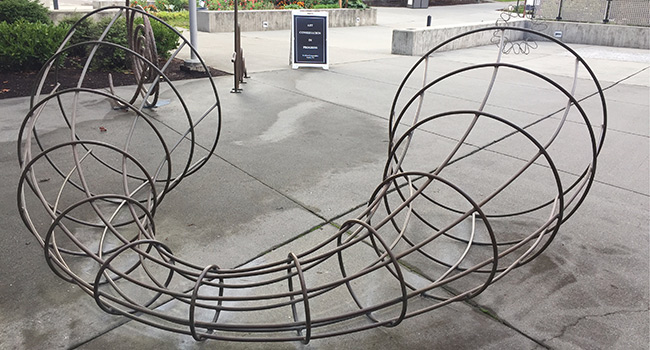 An outdoor plaza with a large metal artwork. Bronze rings and curves create the form of a wormhole curled in half. In the background are more metal sculptures, a sandwich board, buildings and trees.