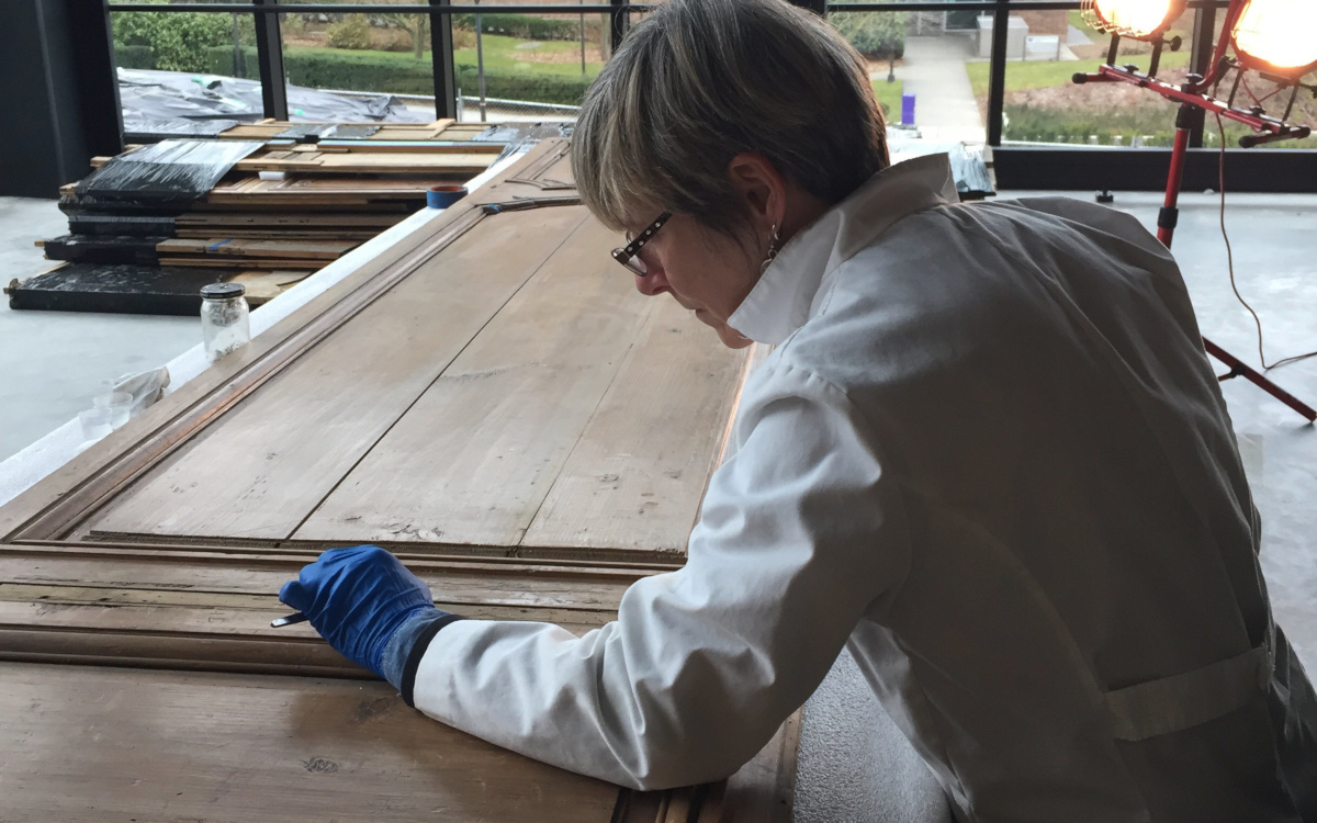 A woman wearing a lab coat and blue nitrile gloves is performiong art conservation treatment on a large carved wooden panel that is laying flat on a table. In the background are work lights and windows.